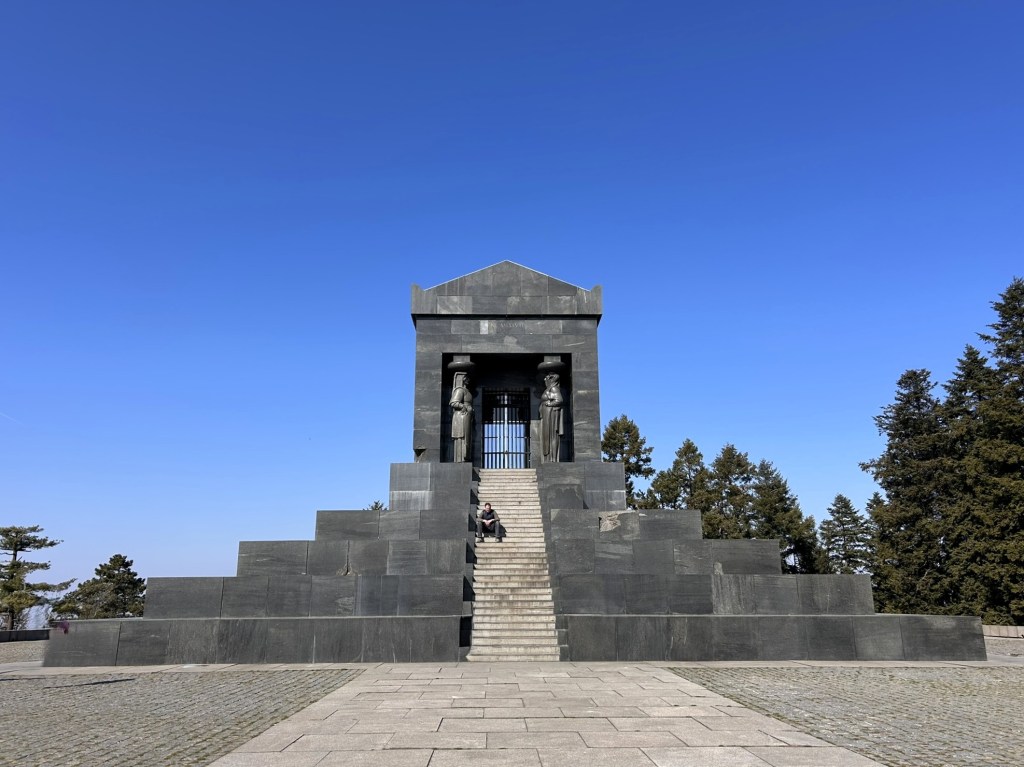 A black stone monument with two statues flanking an entrance, set on a stepped base, against a clear blue sky.