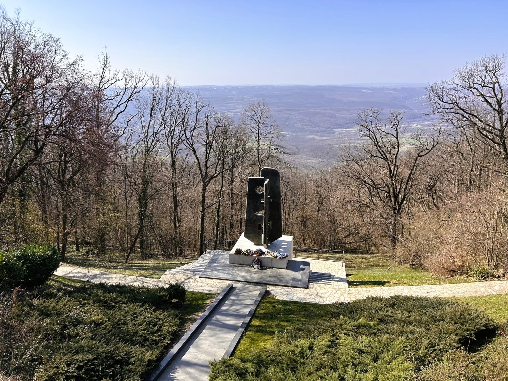 A memorial structure located on a hillside, surrounded by bare trees and a clear blue sky, with a pathway leading to it.