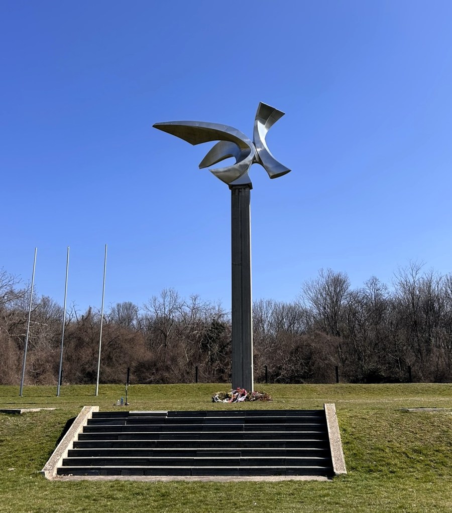 A modern metal sculpture resembling a bird, perched atop a tall pedestal, set against a clear blue sky with trees in the background. Steps lead up to the sculpture, with a small wreath at the base.