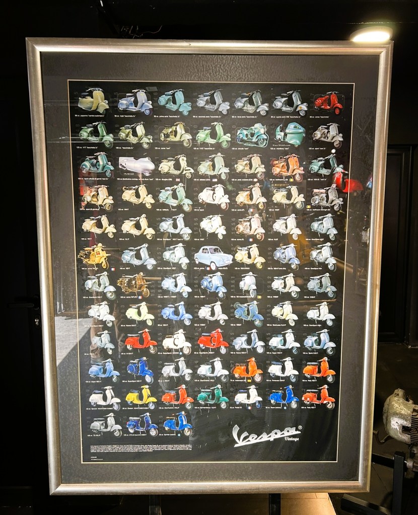 A framed poster displaying various models of Vespa scooters in multiple colours, arranged in a grid format.