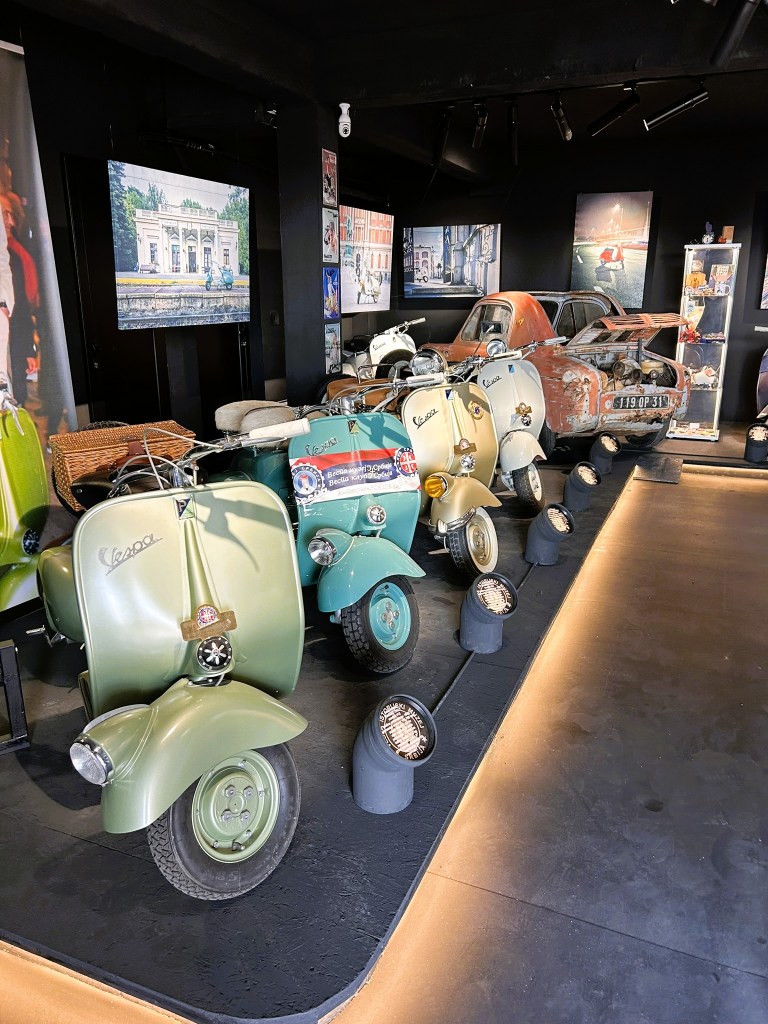 A display of classic Vespa scooters in various shades of green, positioned in an indoor exhibition space. The background features framed photographs and a vintage car, with ambient lighting highlighting the vehicles.