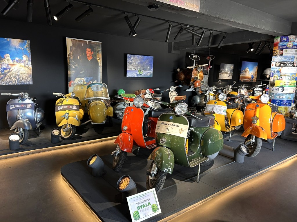 A display of vintage scooters in a museum setting, featuring a variety of brightly coloured Vespas and other models, with photographs and artworks on the walls.