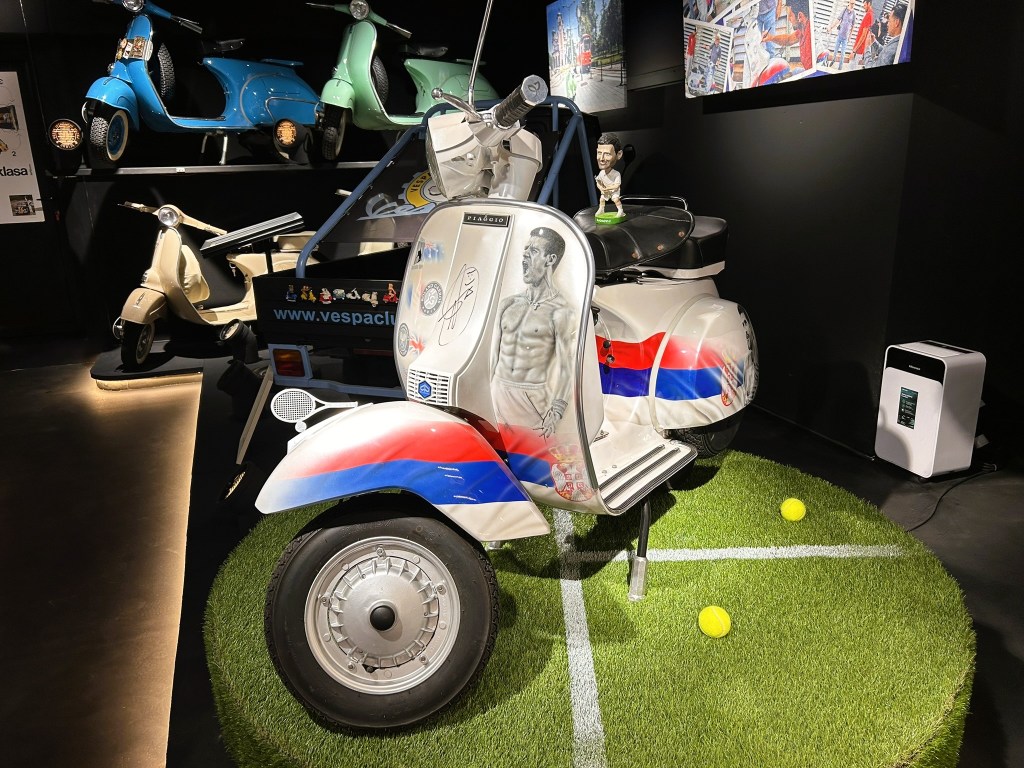 A customised vintage scooter displayed on artificial grass, featuring designs in red, white, and blue, along with a drawing of a muscular figure. Two tennis balls are placed nearby.