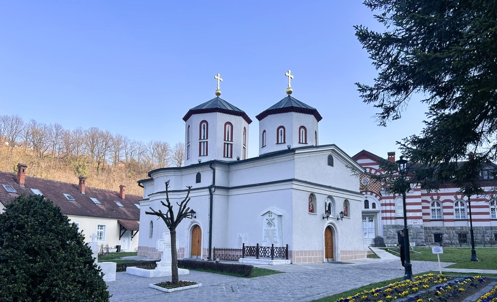 A small white church with two domed towers and gold crosses, set against a clear blue sky. The church is surrounded by greenery and features decorative elements on its façade. In the background, there are buildings with red and white stripes and trees.