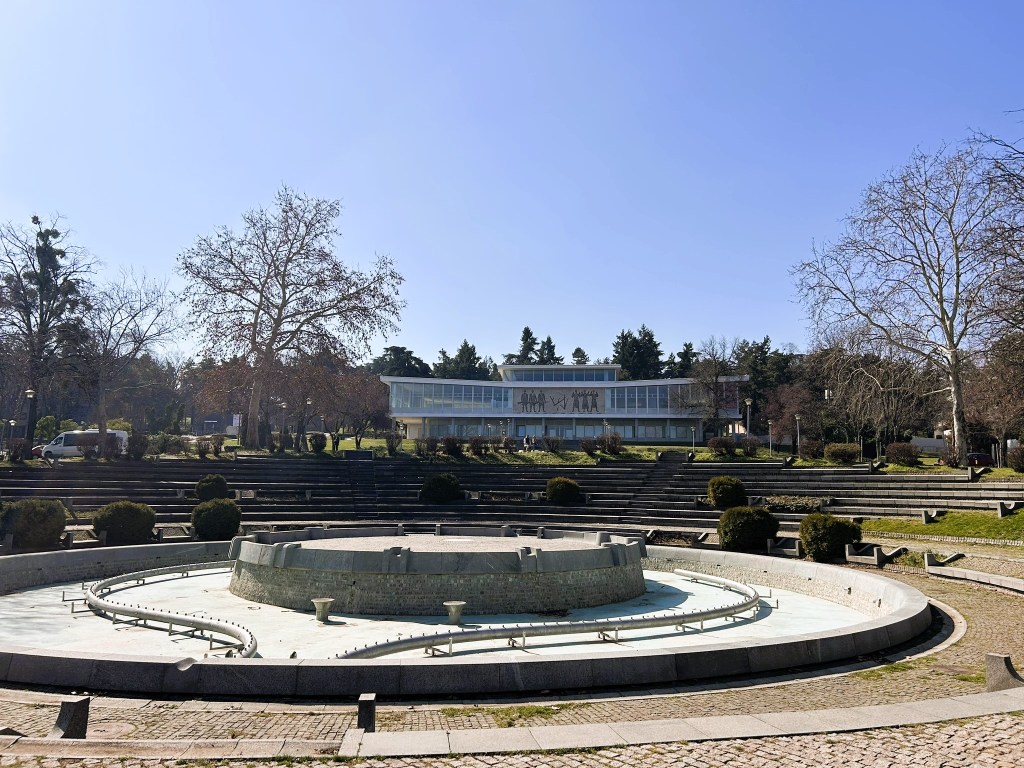 A circular fountain in a park setting, with a modern building in the background and clear blue skies.