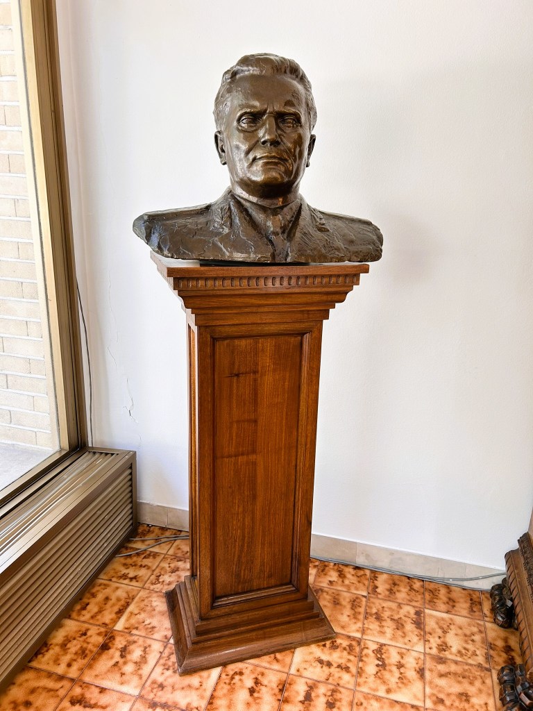 A bronze bust of a man displayed on a wooden pedestal, set against a white wall with a tiled floor.