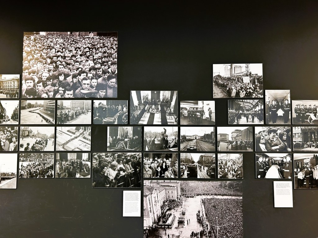 A collage of black and white historical photographs exhibited on a dark wall, depicting crowds, public gatherings, and significant events, with a large image of a dense crowd at the top.