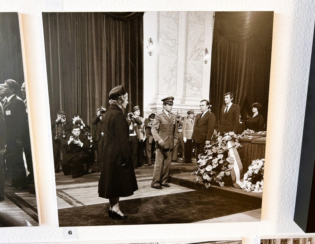 A historical black and white image depicting a solemn ceremony, featuring a woman in a dark coat and hat standing before a military officer in uniform, with several individuals observing and taking photographs in the background.
