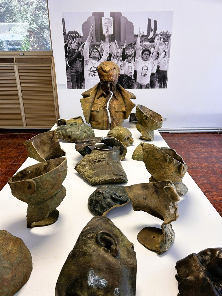 A sculpture display featuring a central bust and several fragmented heads arranged on a table, with a historical black and white photograph of a crowd raising their arms in the background.
