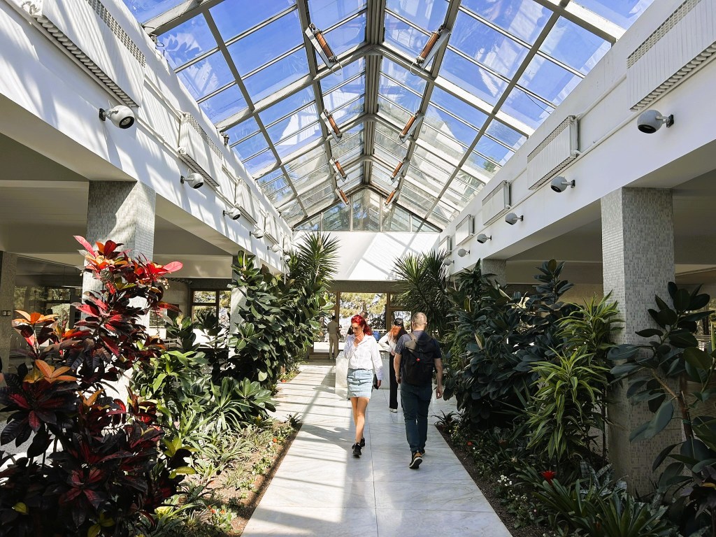 A modern glasshouse interior featuring two people walking along a path surrounded by various tropical plants and flowers under a bright blue sky.