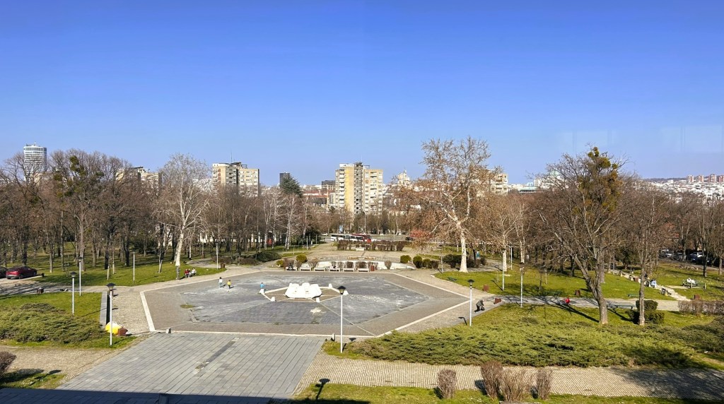 A park view featuring bare trees and a rectangular plaza with a central fountain, surrounded by green grass and pathways, with buildings in the background under a clear blue sky.
