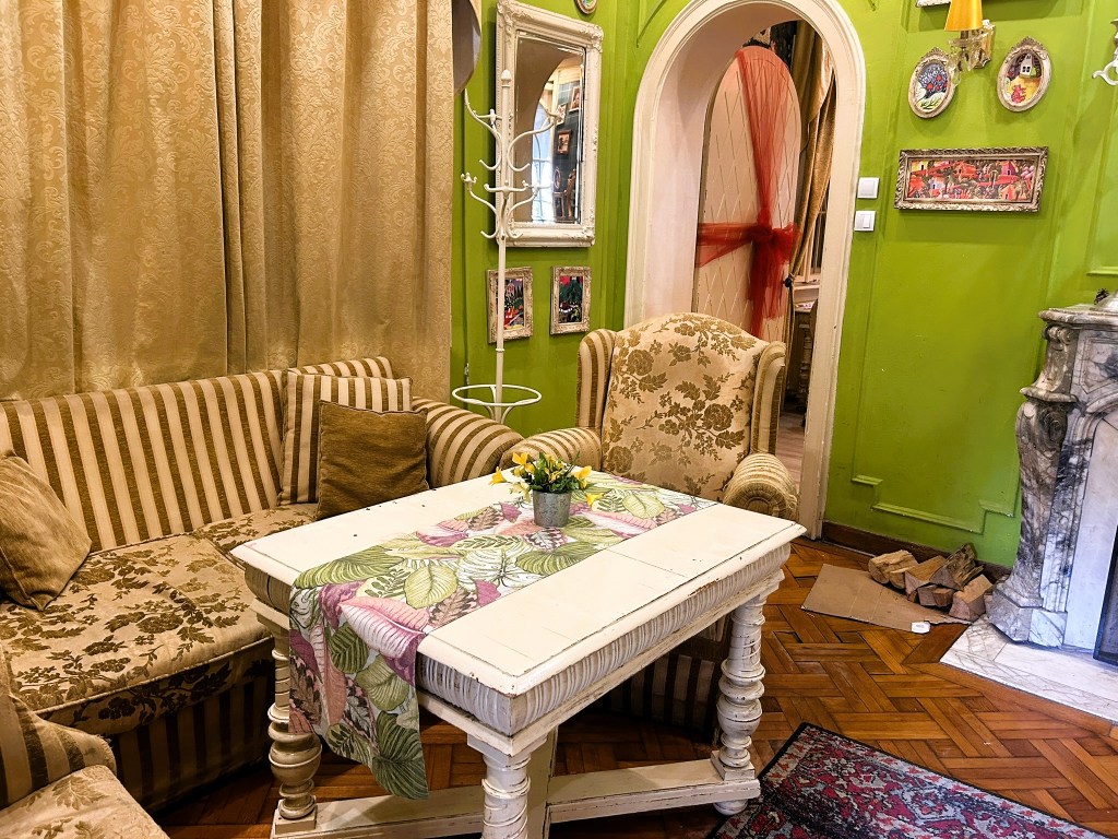 A cosy living room featuring a striped sofa and a floral armchair, with a white wooden table adorned with a colourful tablecloth and a small flower vase. The walls are painted green with decorative frames and curtains in a gold tone.