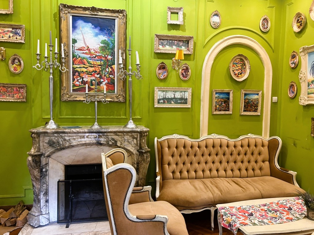 A cosy living room with a bright green wall, featuring a marble fireplace adorned with candelabras. Vintage paintings in ornate frames decorate the wall, along with a golden-framed artwork above the fireplace. A plush, tufted beige sofa and an armchair complete the inviting atmosphere.
