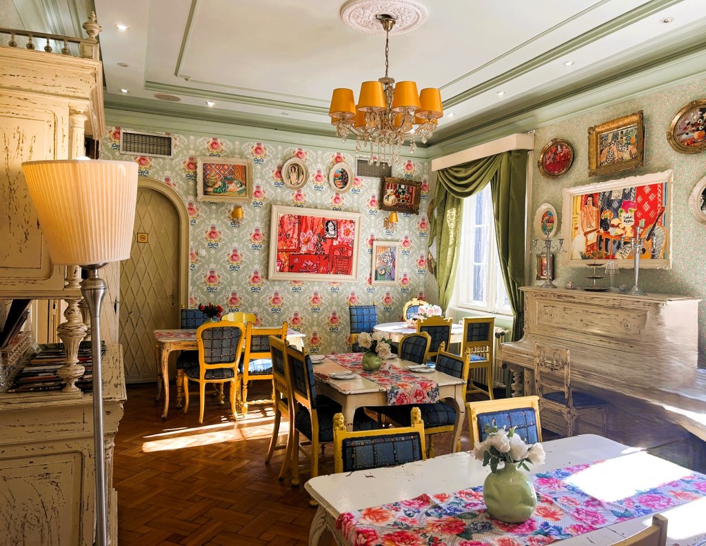 A beautifully decorated dining room featuring floral wallpaper, ornate furniture, and an eclectic mix of artwork on the walls. There are wooden tables with floral tablecloths and colourful chairs. A piano and a lamp add to the charm of the space.