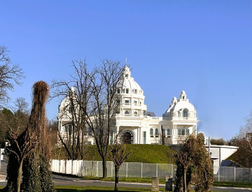 A large, ornate white building with multiple domes and architectural details, set against a clear blue sky. In the foreground, there are several bare trees and sculptures resembling topiary.