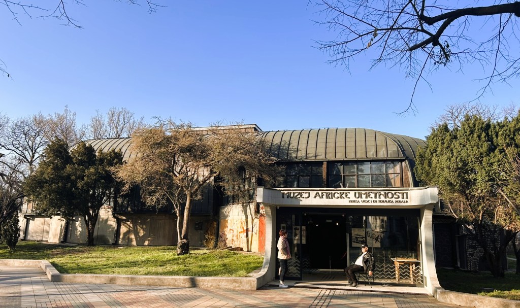 Exterior view of the African Art Museum, featuring a modern architectural design with a curved roof and large windows. Two people stand outside the entrance amidst greenery under a clear blue sky.