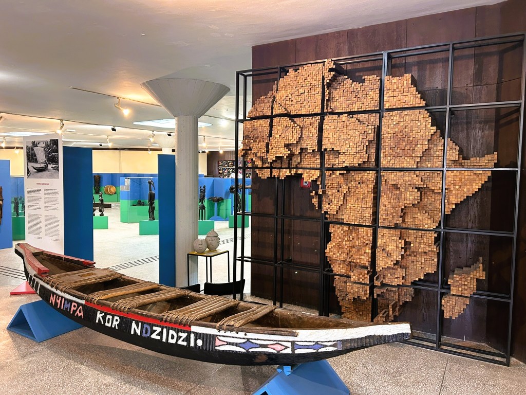 An intricately decorated wooden canoe displayed in an exhibition space, with a large, artistic representation of the African continent made from small wooden blocks in the background.