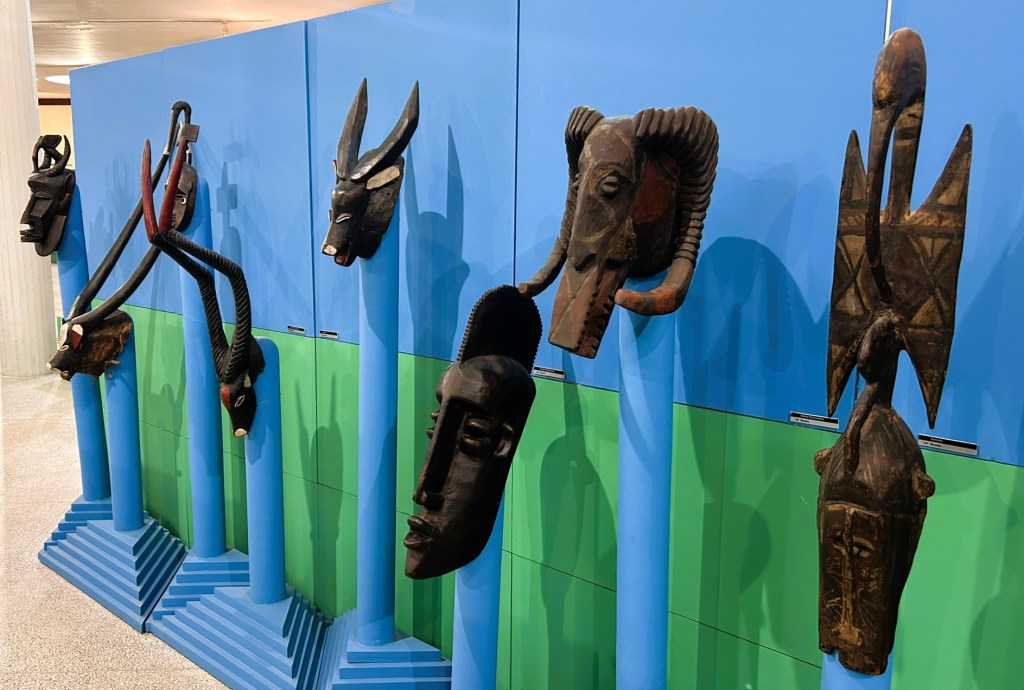 A display of various traditional masks mounted on blue poles, featuring intricate designs and carvings, set against a green background.