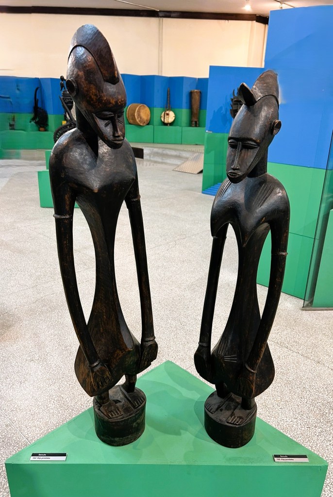 Two wooden sculptures depicting abstract human figures, displayed on a green pedestal in a museum setting.