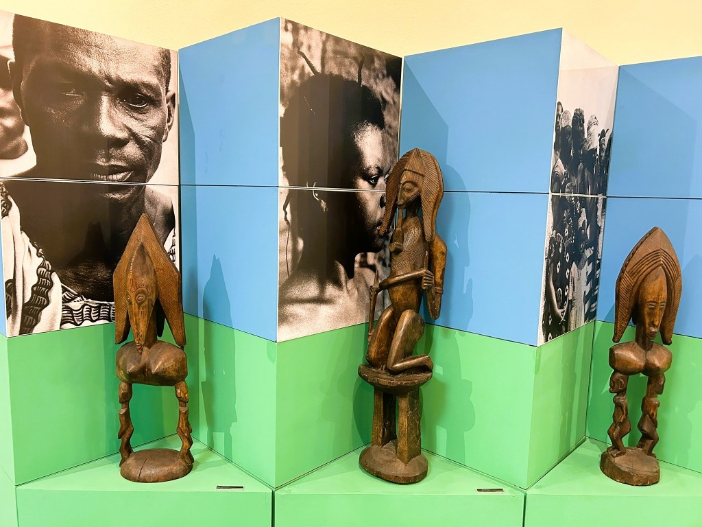 Two wooden sculptures on display in an exhibition, surrounded by colourful panels featuring black and white photographs of people.