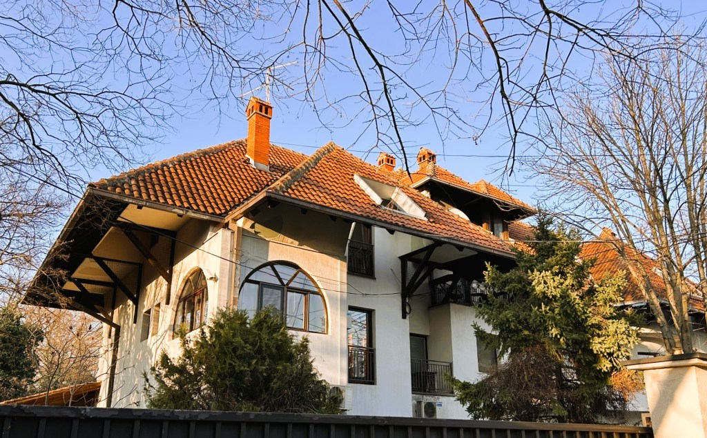 View of a large, white house with a red tiled roof, surrounded by trees and clear blue sky.