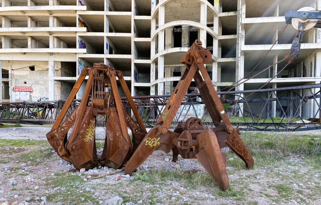 Rusty mechanical claws used for construction, positioned in front of an abandoned building with graffiti on its concrete walls.