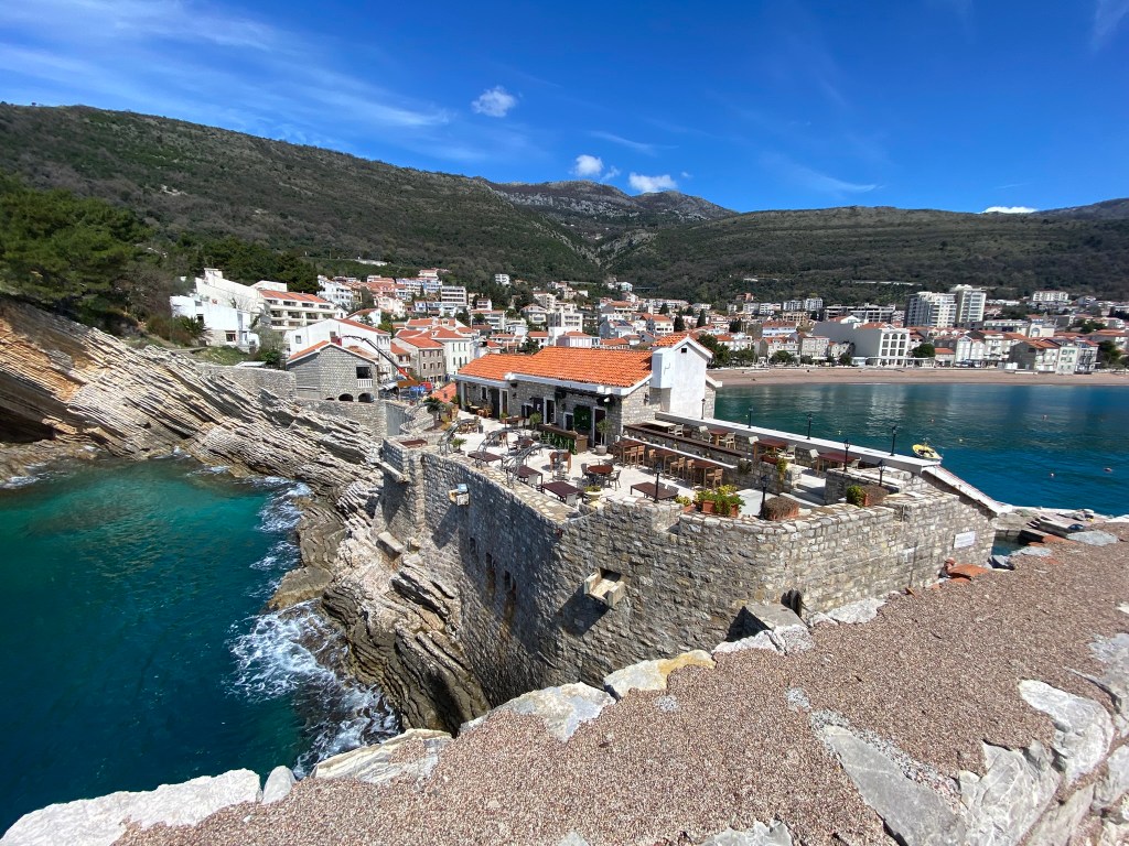 A coastal view showing a restaurant built on rocky cliffs, overlooking turquoise waters with a backdrop of hills and a town with residential buildings.