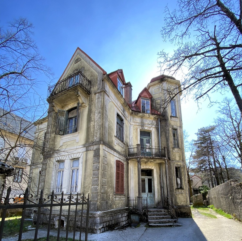 An old, weathered two-storey house with a red-tiled roof, featuring a balcony and multiple windows, surrounded by bare trees and blue sky.