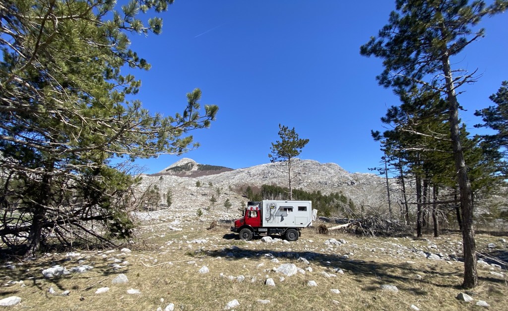 A red and white off-road vehicle parked in a rocky clearing surrounded by trees and mountains under a clear blue sky.