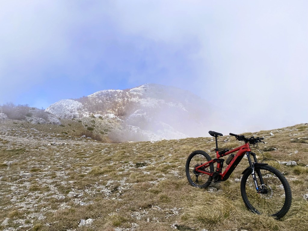 A red mountain bike resting on grassy terrain with rocky outcrops and a misty mountain backdrop under a blue sky.