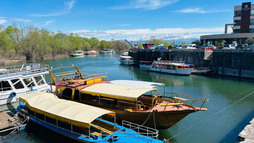 A view of a calm waterway with several boats moored, including a brightly coloured yellow and blue boat. In the background, there are trees and a building, with a clear blue sky and mountains visible in the distance.