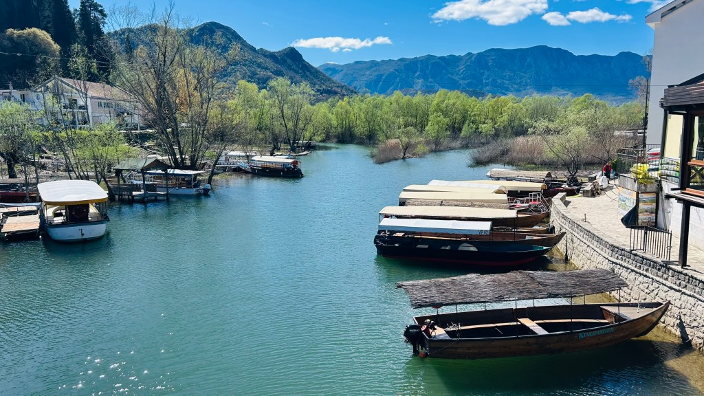 A scenic view of a river with several boats docked along the shore, surrounded by green trees and mountains in the background under a blue sky.