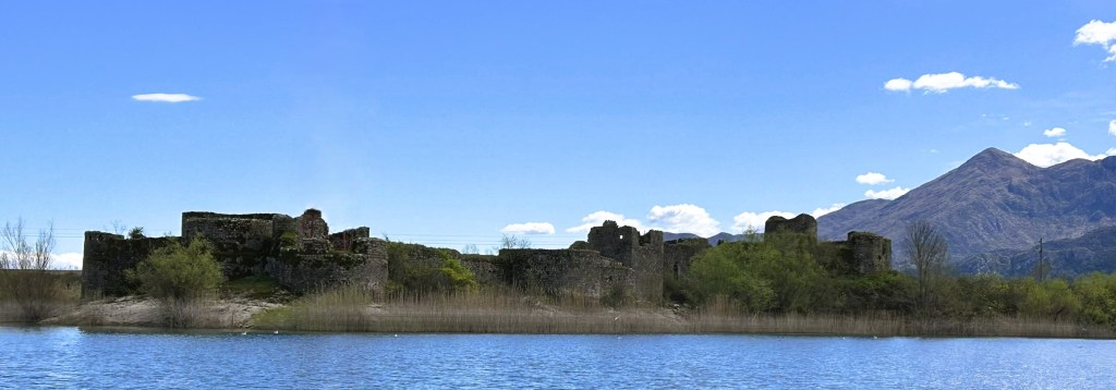 A scenic view of historical ruins beside a calm lake, with mountains in the background under a clear blue sky.
