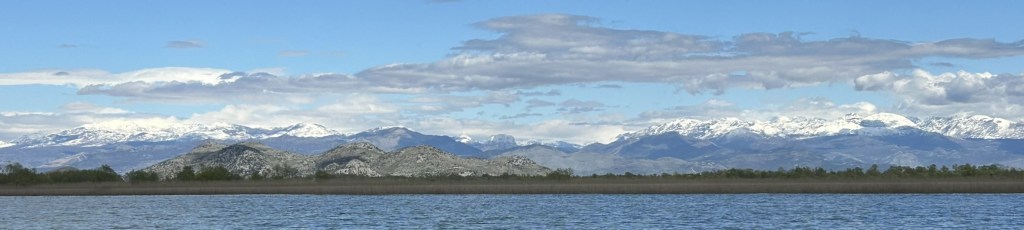 A panoramic view of snow-capped mountains under a blue sky with scattered clouds, framed by a calm lake and a line of greenery in the foreground.