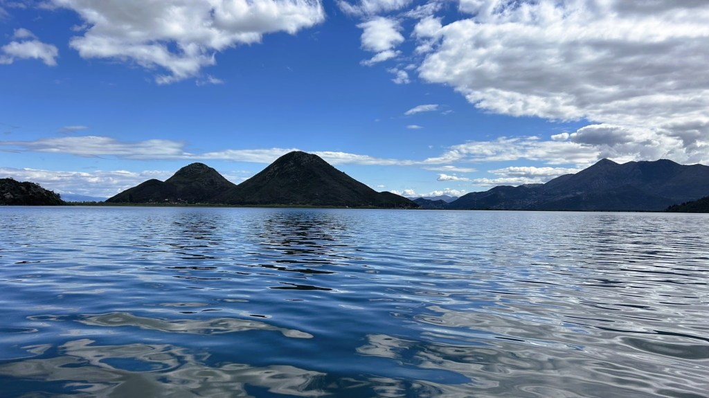 A serene view of calm water with mountain peaks in the background under a blue sky with scattered clouds.