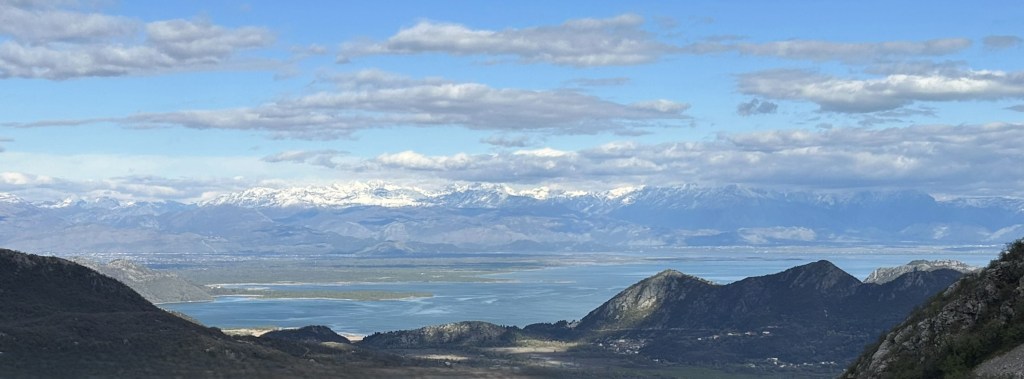 Panoramic view of a mountainous landscape with snow-capped peaks, a large water body, and fluffy clouds in a blue sky.