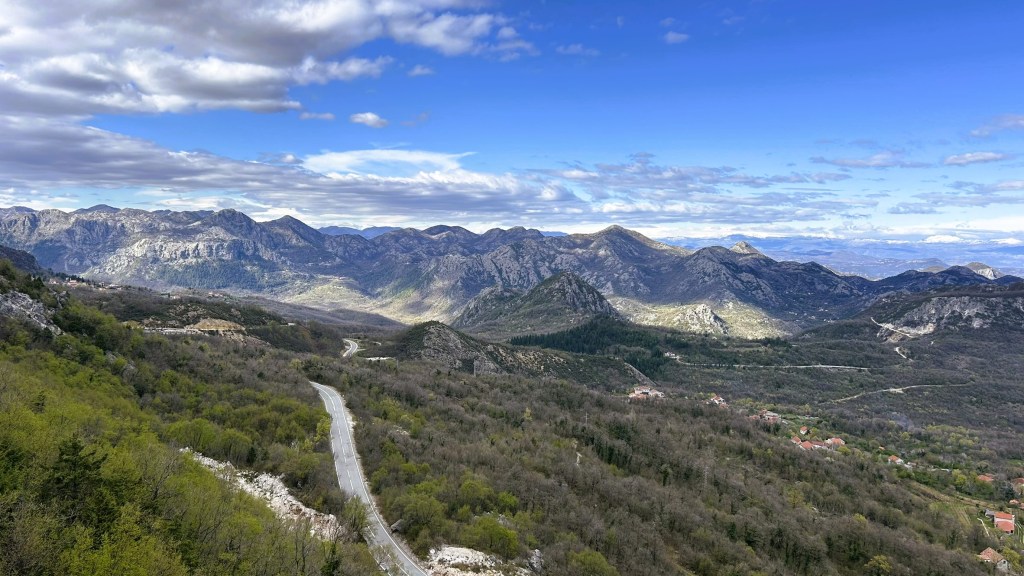 Panoramic view of mountainous landscape with winding road and lush green hills under a partly cloudy sky.