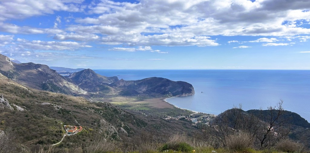 A panoramic view of a coastal landscape featuring mountains, a sweeping shoreline, and a clear blue sea under a partly cloudy sky.