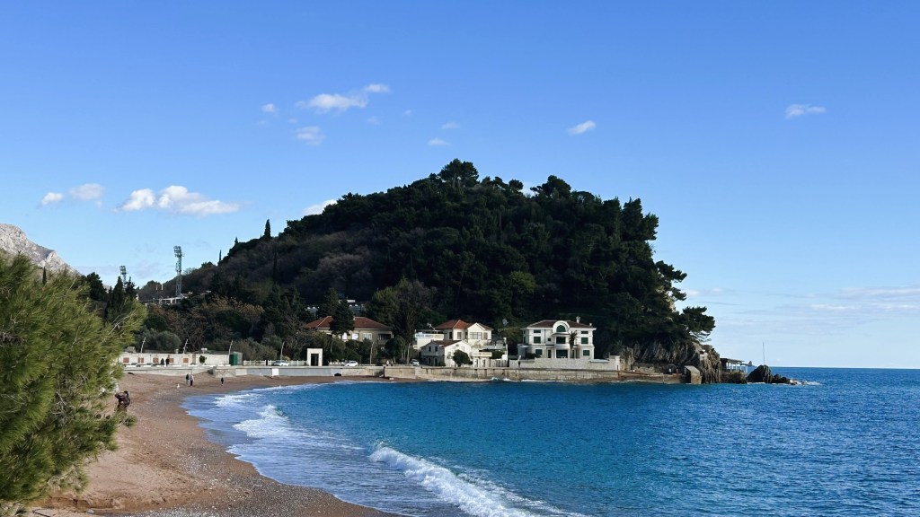 View of a beach with calm blue waters, featuring a rocky hill covered in greenery and several buildings along the shore under a clear blue sky.
