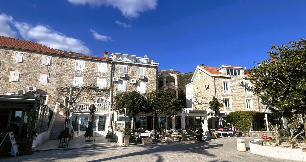 A scenic view of a stone-paved square surrounded by traditional stone buildings with white shutters, featuring outdoor seating areas and trees under a clear blue sky.