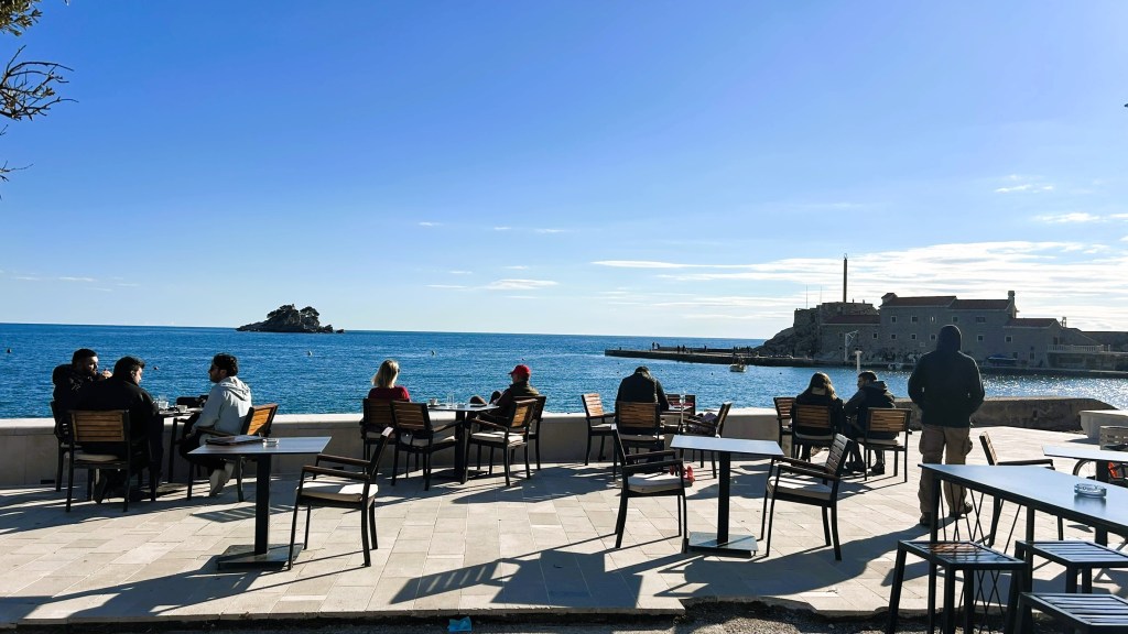 People seated at outdoor tables by the seaside, enjoying views of the water and a distant rocky island under a clear blue sky.