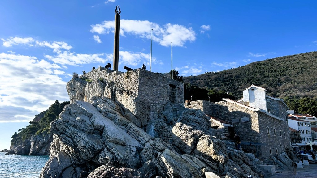 A coastal stone fortress situated on rocky terrain, overlooking the sea, with a tall metal pole and several flags against a backdrop of hills and a blue sky.
