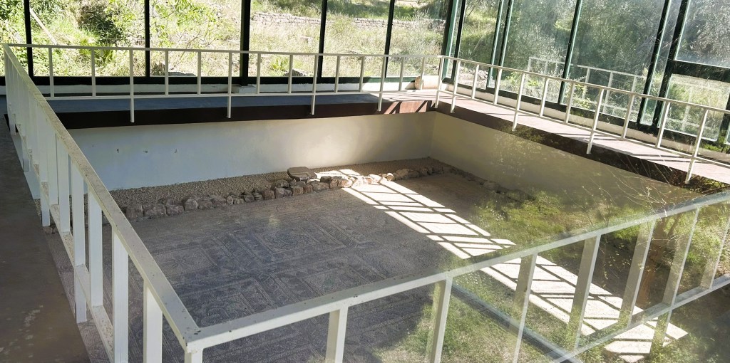 An archaeological site featuring an exposed mosaic floor enclosed within a glass structure, surrounded by greenery.