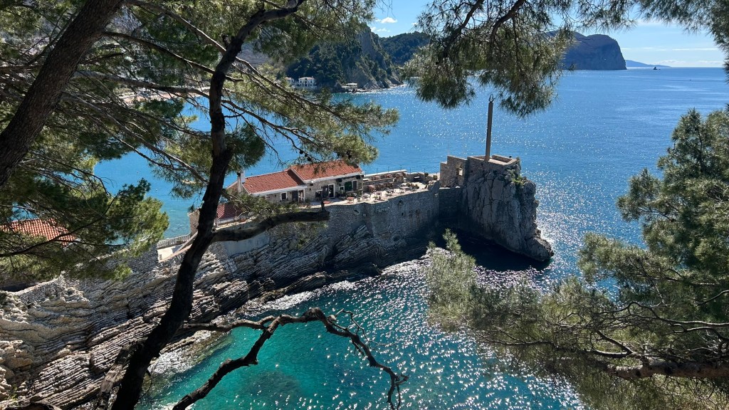 A scenic view of a coastal structure on a rocky outcrop surrounded by clear turquoise water, with trees framing the image and distant hills in the background.