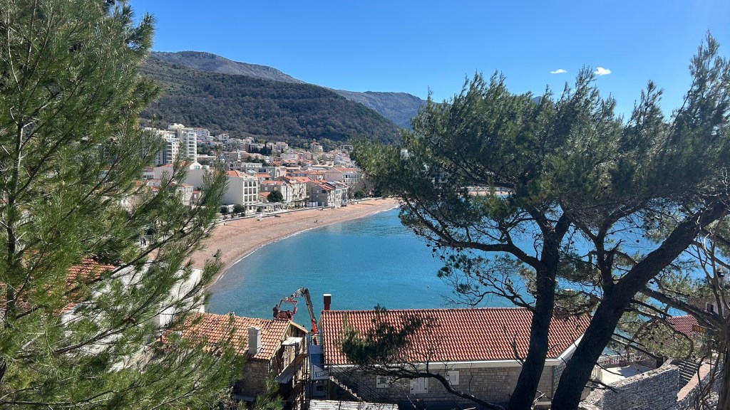 A scenic view of a coastline with a sandy beach, framed by green trees and backed by mountains. The shoreline features a small town with buildings along the beach, under a clear blue sky.