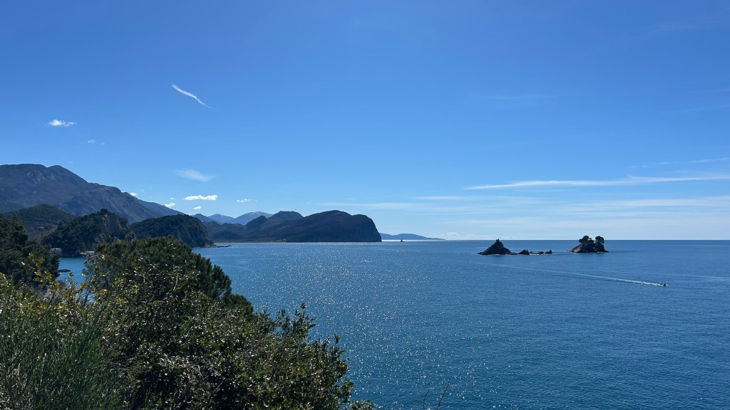 View of a serene coastal landscape featuring a calm sea, distant mountains, and small rocky islands under a clear blue sky.