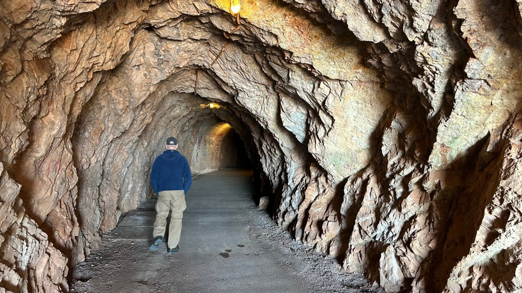 A person walking through a rocky tunnel with uneven walls, illuminated by overhead lights.
