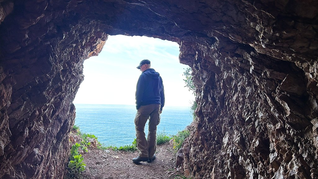 A person standing in a cave-like opening, looking out at the ocean and sky.