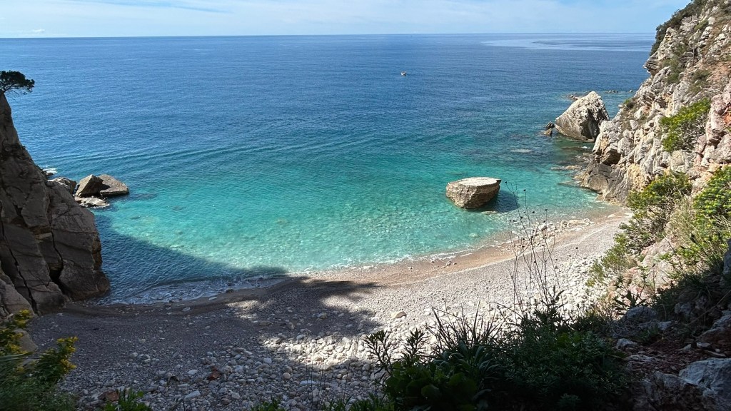A tranquil coastal scene featuring clear turquoise waters, rocky cliffs, and a small pebble beach.
