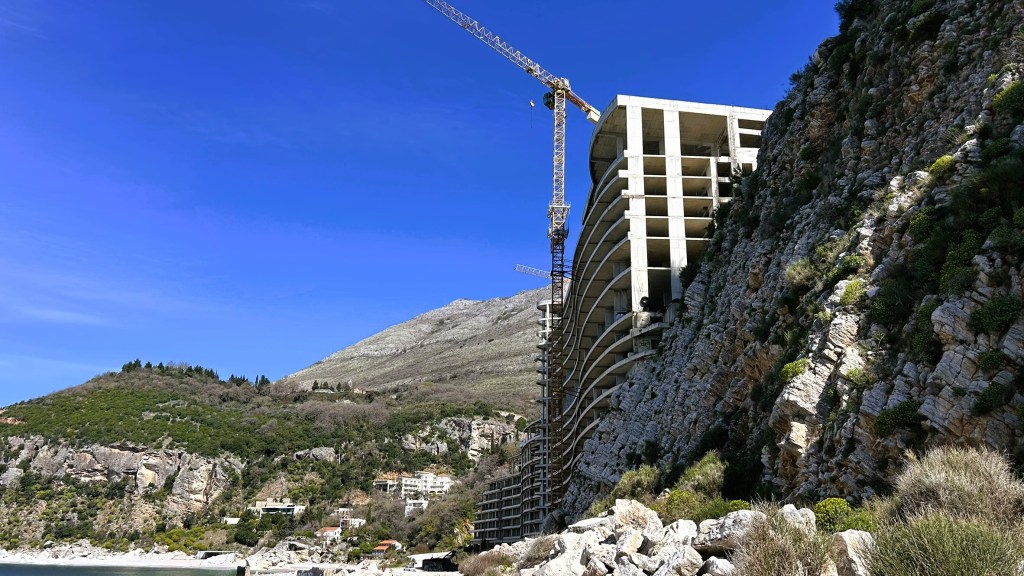 Construction site of a large building next to a rocky coastline, with a crane in the foreground and a mountain in the background under a clear blue sky.